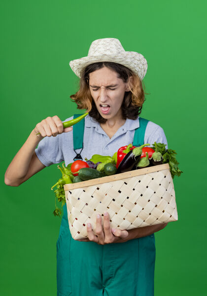 男人年轻的园丁穿着连体衣戴着帽子拿着装满新鲜蔬菜的箱子辣椒蔬菜连身衣