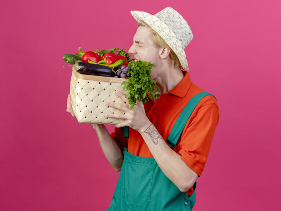 蔬菜年轻的园丁穿着连体衣戴着帽子拿着装满蔬菜的箱子持有连身衣园丁