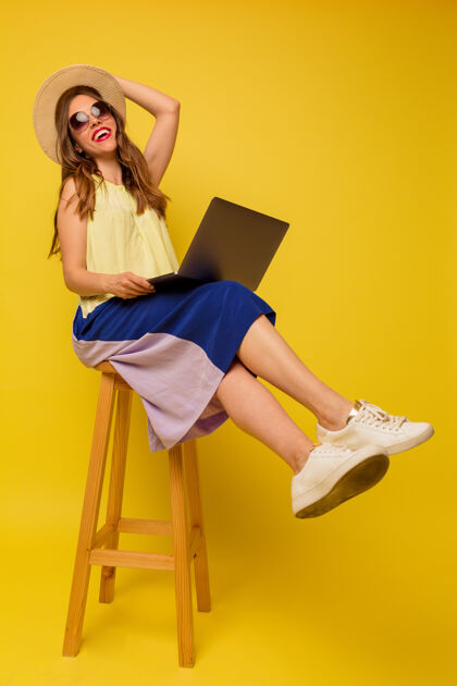 积极穿着鲜艳夏装的轻松女孩拿着笔记本电脑坐在隔离墙上的椅子上工作乐观民族快乐