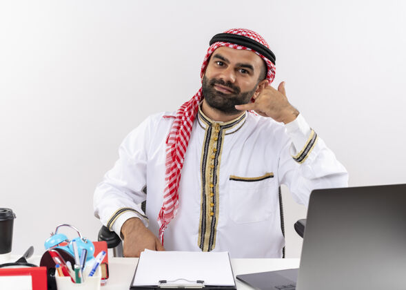 电脑身着传统服装的阿拉伯商人坐在桌旁 用笔记本电脑打电话给我 微笑着在办公室工作电话阿拉伯语笔记本电脑