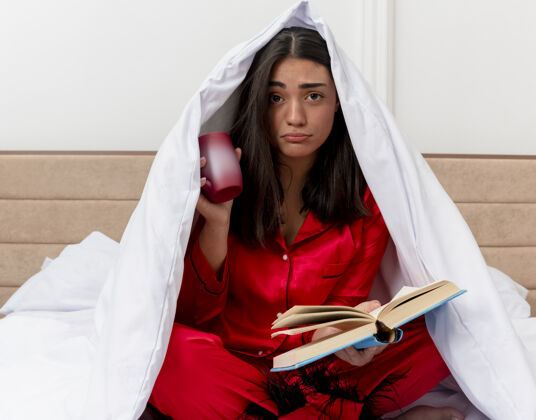 床穿着红色睡衣的年轻美女坐在床上 裹着毛毯 端着咖啡和书 在灯光背景下看着卧室里表情悲伤的相机相机红色表情
