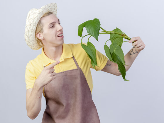 手指年轻的园丁 穿着连体衣 戴着帽子 拿着一株植物 用食指指着它 站在白色的背景上 满脸笑容帽子植物快乐