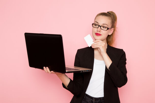 女职员身穿黑色夹克 手持白色卡片 在粉色墙上使用笔记本电脑的女上班族正面图微笑工人人