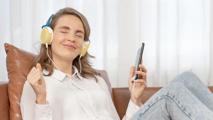 空间生活方式美丽可爱的女孩女人感觉快乐享受在沙发上用耳机听音乐使用音乐女孩