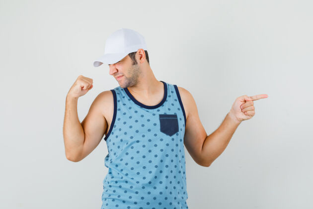 衬衫年轻人指着旁边 在蓝色的单打 帽子和看起来高兴的赢家的姿态秀现代胡须