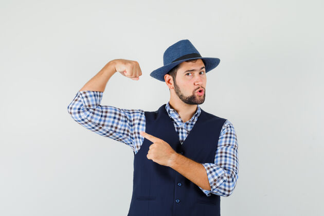 胡茬年轻人指着穿着衬衫 背心 帽子的手臂肌肉 看上去很强壮前视图手臂男人站立