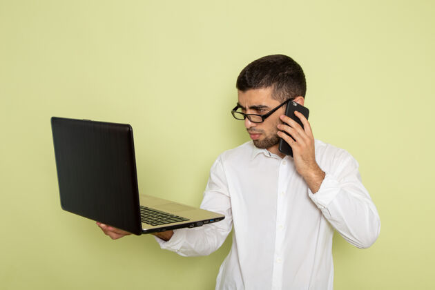 工人身穿白衬衫的男办公室工作人员在绿色墙上打电话和使用笔记本电脑的正面图工作衬衫电话