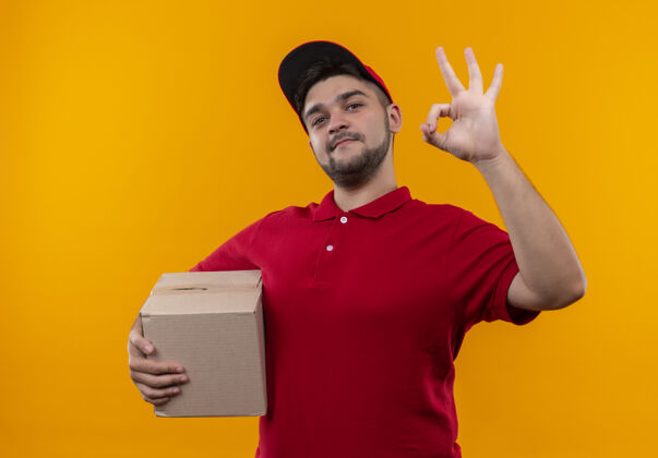 站着年轻的送货员穿着红色制服 戴着帽子 手里拿着盒子 微笑着 自信地展示着ok标志包装帽子自信