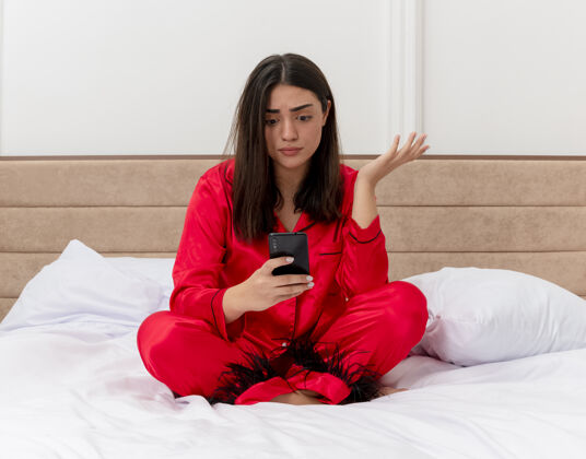 漂亮穿着红色睡衣的年轻漂亮女人坐在床上用智能手机看着卧室内部灯光背景下的困惑和不快坐着床困惑