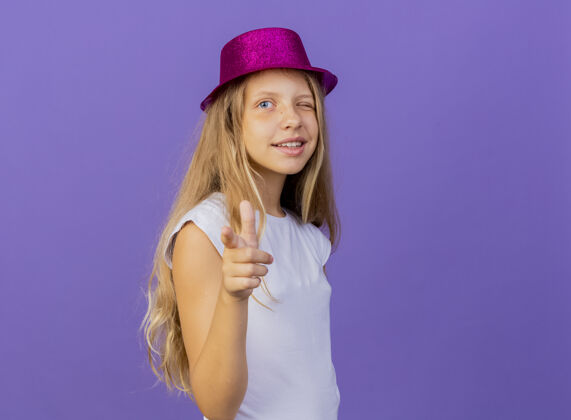 手指戴着节日礼帽的漂亮小女孩用食指指着镜头微笑着眨眼 紫色背景下站着生日派对的概念生日小眨眼