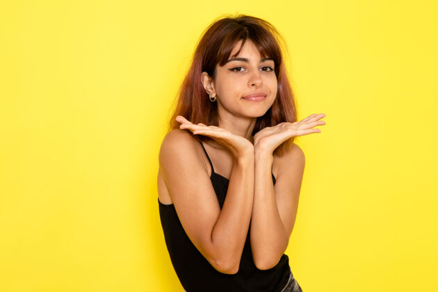 性感身穿黑色衬衫的年轻女性在浅黄色墙壁上摆出可爱表情的正面图模特衬衫情感
