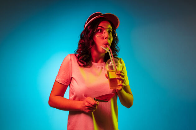 波浪戴着帽子拿着糖果和饮料在时髦的蓝色霓虹灯工作室的年轻女子令人心寒派对表情饮料