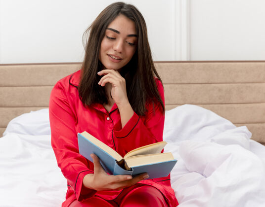 室内穿着红色睡衣的年轻漂亮女人坐在床上 在卧室的室内灯光背景下愉快而积极地看书阅读年轻床