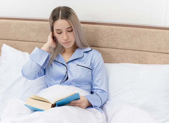 漂亮穿着蓝色睡衣的年轻美女坐在床上 在卧室的室内灯光背景下看书床年轻卧室