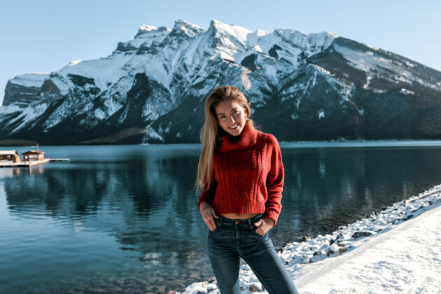 红唇美丽的女士站在湖边的沙滩上 带着白色的微笑山上覆盖着雪穿着红色的针织毛衣和蓝色的牛仔裤金色的长发 没有化妆现代爱情时尚
