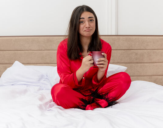 美丽穿着红色睡衣的不快乐的年轻漂亮女人坐在床上 端着一杯咖啡 看着相机 在卧室的室内灯光背景下 带着悲伤的表情床表情睡衣
