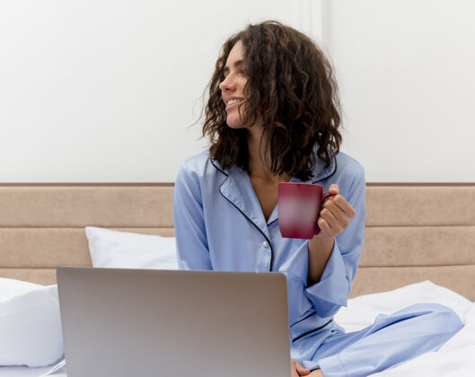 坐着穿着蓝色睡衣的有趣年轻漂亮女人坐在床上喝着咖啡在笔记本电脑上工作愉快而积极地看着卧室内部的灯光背景睡衣搞笑快乐
