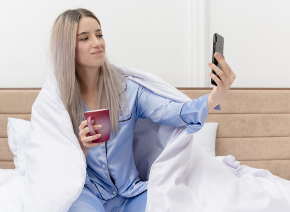 咖啡穿着蓝色睡衣的年轻美女坐在床上 喝着咖啡 在明亮的背景下 用智能手机在卧室里快乐而积极地自拍室内床睡衣