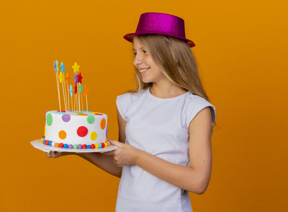 生日戴着节日礼帽的漂亮小女孩拿着生日蛋糕 面带微笑 站在橙色背景下的生日派对概念微笑橙色站着