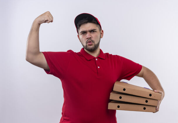 制服身穿红色制服 头戴鸭舌帽的年轻送货员举着披萨盒 举起拳头 表情严肃地展示二头肌 赢家概念二头肌年轻赢家