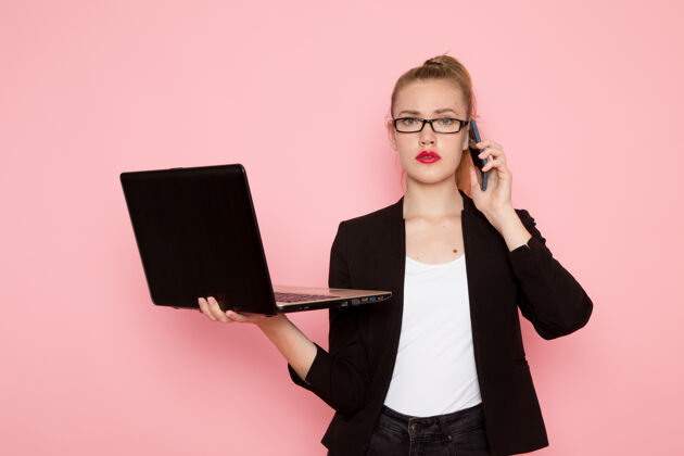 业务穿着黑色夹克的女上班族正对着淡粉色的墙上拿着和使用笔记本电脑说话工人持有女商人