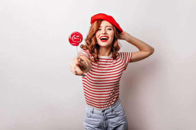 肖像穿着红色贝雷帽吃糖果的可爱女孩姜黄色头发和棒棒糖摆姿势的法国女士姜棒棒糖甜点