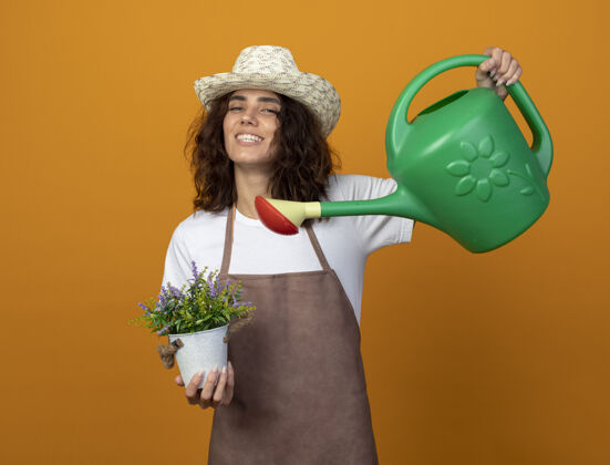休闲微笑的年轻女园丁穿着制服戴着园艺帽在花盆里浇花用浇水罐隔离在橙色的墙上女人年轻花园