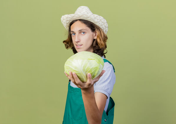 男人年轻的园丁穿着连体衣 戴着帽子 手里拿着卷心菜 站在绿色的背景下 自信地微笑着看着镜头绿色抱着自信