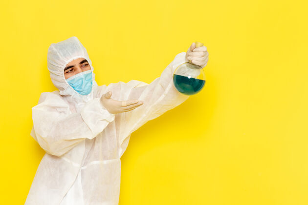 手持穿着特殊防护服的男科学工作者的正面图 黄色墙上拿着装有绿色溶液的烧瓶医疗正面危险