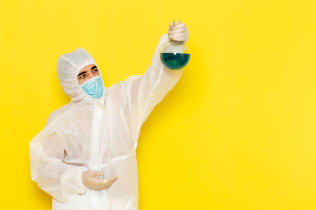 男性穿着特殊防护服的男性科学工作者的正面图 黄色墙壁上拿着装有绿色溶液的烧瓶正面医学医生