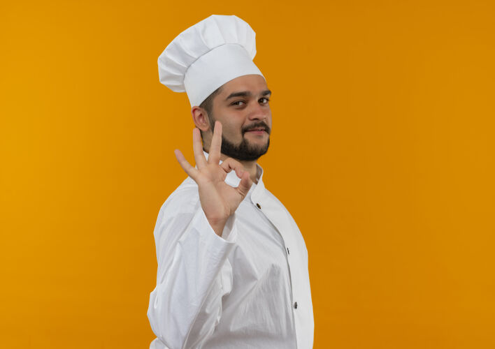 好身着厨师制服的年轻男厨师站在侧视图中 并在橙色空间上孤立地做着“ok”标志复制男性年轻