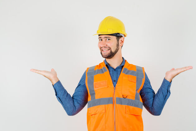 年轻人年轻的建筑工人在衬衫 背心 头盔上做着比例尺的手势 看上去很开朗 前视图工作车间勤杂工
