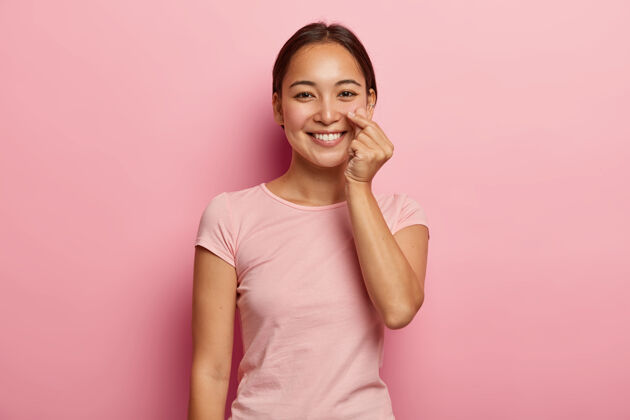 牙关身体语言概念快乐的亚洲女人做韩语手势 表达爱 做喜欢的手势 微笑温柔 穿着休闲服 隔离在粉红色的墙上单色穿孔积极浪漫