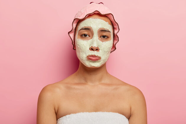 清洁心烦意乱的韩国年轻女性有不满意的悲伤表情 接受美容治疗 不高兴有皱纹和有问题的皮肤 脸上有化妆面膜 头戴防水淋浴帽治疗悲伤模特