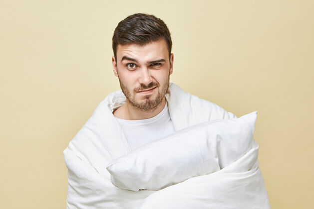 疲倦照片中 沮丧的年轻人没有刮胡子 感觉压力很大 要早起 裹在白色柔软的毯子里 手里拿着枕头 表情愤怒床上用品概念枕头卧室舒适