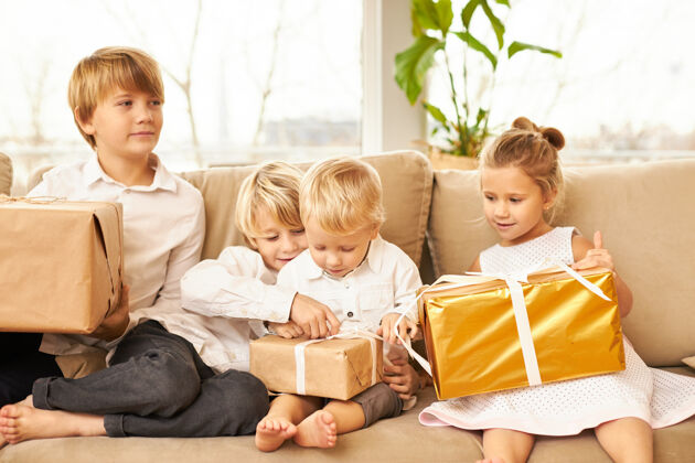 孩子四个白人孩子穿着同样的白衬衫 没有袜子 坐在客厅的沙发上 迫不及待地打开装有新年礼物的盒子 面带微笑 脸上洋溢着喜悦和兴奋的表情小童年圣诞老人