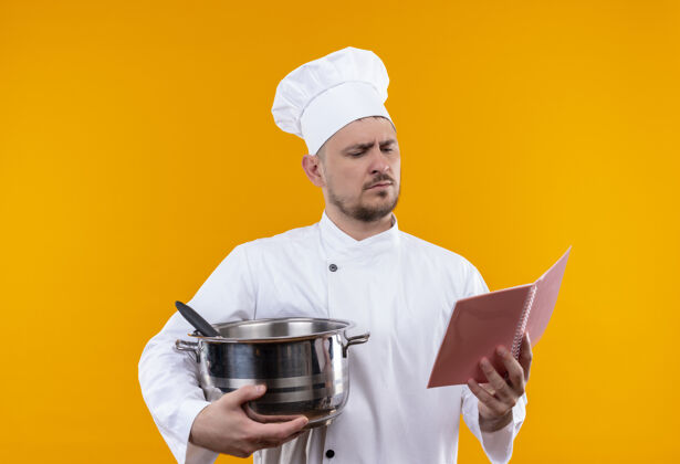 年轻年轻帅气的厨师穿着厨师制服 拿着锅炉和记事本 看着孤立的橙色空间上的记事本厨师帅气拿着