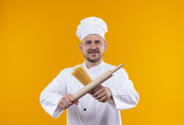 拿着身着厨师制服的年轻帅哥面带微笑 手里拿着意大利面和擀面杖 孤立地站在橙色的空间里意大利面橙色厨师