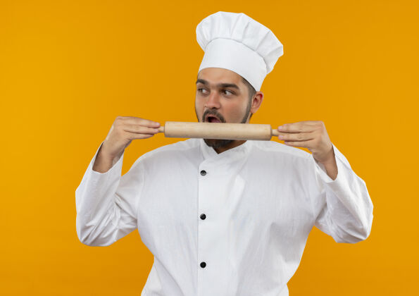 抱着年轻的男厨师穿着厨师制服 拿着擀面杖 试图咬着 看着旁边孤立的橘子空间看别针滚动