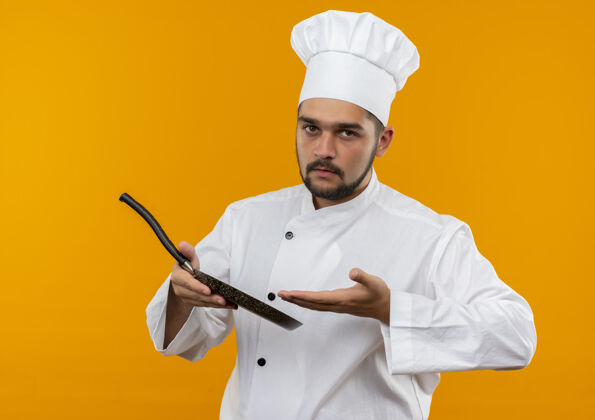 指着身着厨师制服的年轻男厨师手持并指着隔离在橙色空间的煎锅制服拿着橙色