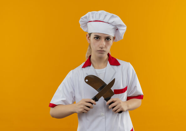 制服年轻漂亮的厨师穿着厨师制服 拿着菜刀和菜刀在橙色的空间里做着“不”的手势手势不愉快切肉刀