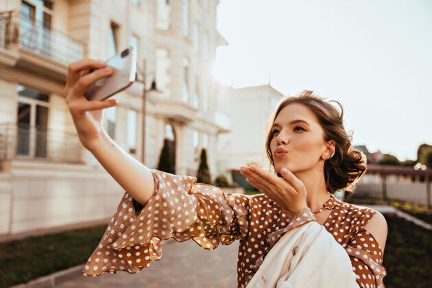 模特优雅的白人年轻女子在秋日用手机自拍身着棕色衣服的迷人女模特送上空中接吻的户外镜头街道脸年轻