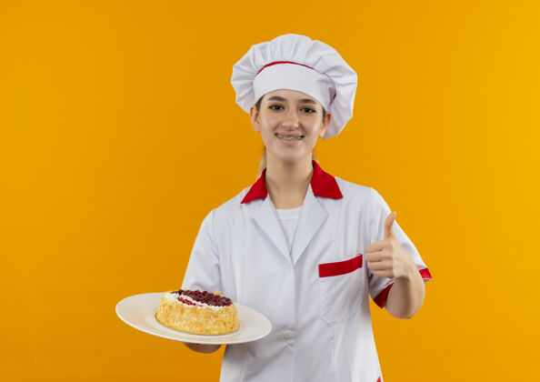 蛋糕微笑着的年轻漂亮的厨师 穿着厨师制服 戴着牙套 拿着一盘蛋糕 在橙色的空间里孤立地竖起大拇指展示拿着厨师
