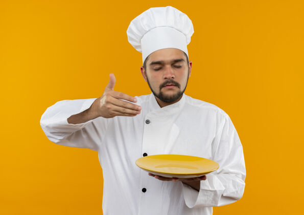 嗅年轻的男厨师穿着厨师制服 拿着盘子 用手嗅着空气 闭着眼睛孤立在橙色的空间里空气盘子年轻