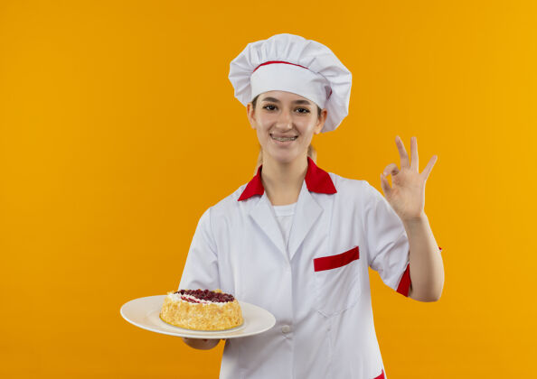 招牌微笑着的年轻漂亮厨师身着厨师制服 戴着牙套 拿着一盘蛋糕 在橙色的空间里隔离着“一切正常”的标志好橘子制服