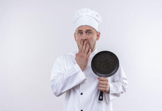 制服惊讶的年轻帅哥厨师穿着厨师制服拿着煎锅把手放在嘴上隔离在空白处嘴惊喜手