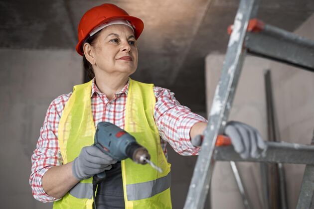 头盔笑眯眯的女建筑工人带着钻工作背心行业