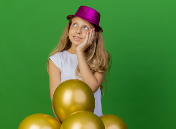 生日戴着节日礼帽的漂亮小女孩和一群巴隆人面带微笑 一边思考 绿色背景下的生日派对概念思想帽子束