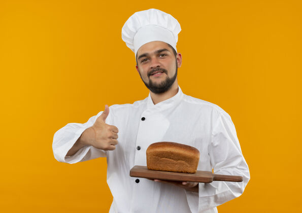 展示身着厨师制服 面带微笑的年轻男厨师手拿着放着面包的砧板 在橙色的空间里孤立地竖起大拇指拇指切割厨师