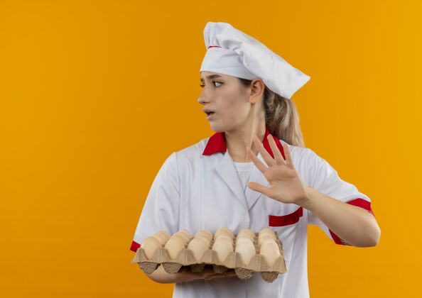 空间年轻漂亮的厨师穿着厨师制服 手里拿着一盒鸡蛋 看着一边 在橙色的空间里做着不孤立的手势厨师漂亮手势